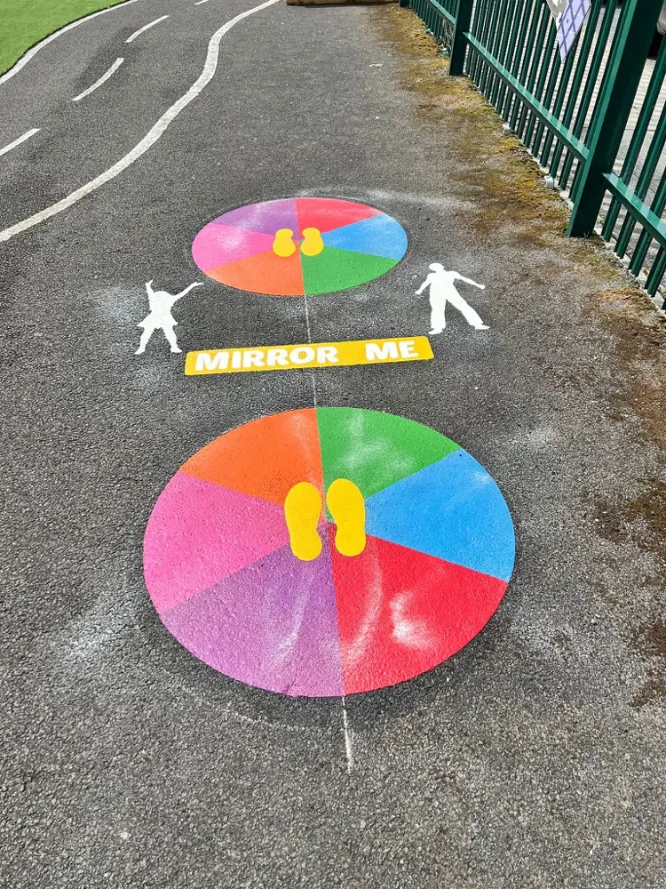 Two colour-wheel Mirror Me Fitness discs with footprints and a MIRROR ME label on a fenced tarmac path