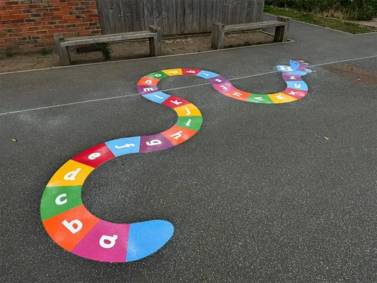 Thermoplastic alphabet snake with rainbow segments lettered a to z, winding across playground tarmac by two wooden benches