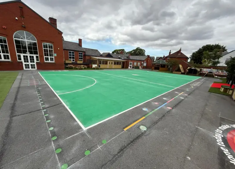 Green netball court with white line markings and coloured dot trails on a playground beside red-brick buildings and play huts