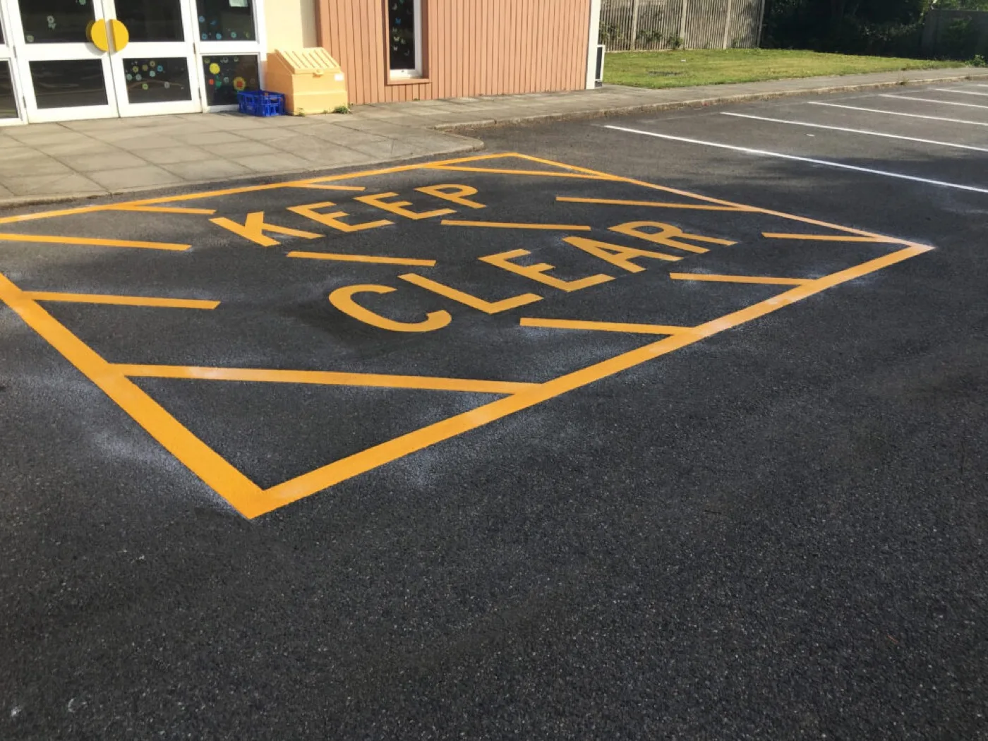 Yellow KEEP CLEAR box marking with a hatched border laid on dark tarmac outside a school entrance