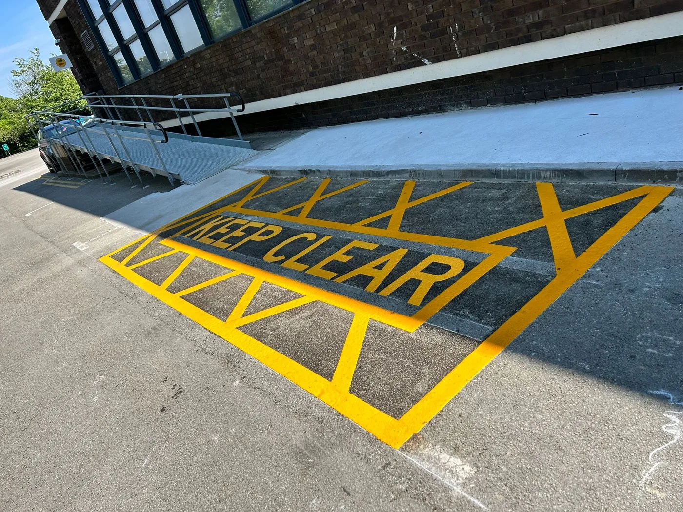 Freshly painted yellow KEEP CLEAR hatched box across an access ramp and lowered kerb beside a building