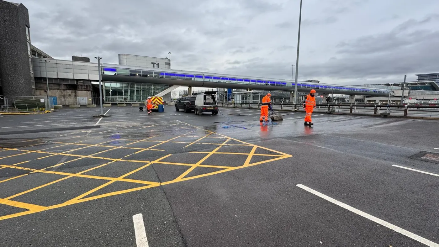 Crew in high-vis using water jetting gear to lift faded yellow box-junction hatching from a tarmac forecourt