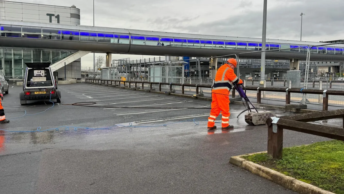 Worker planing off bay markings beside a van-mounted power unit, trailing a hose across the wet car park