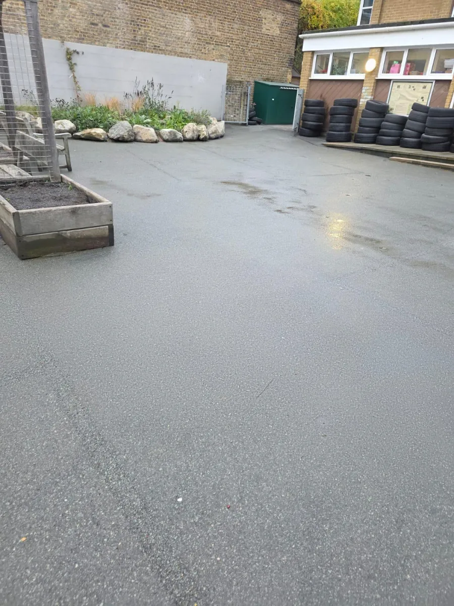 Empty playground surface stripped back to bare grey tarmac beside raised beds and a stack of tyres