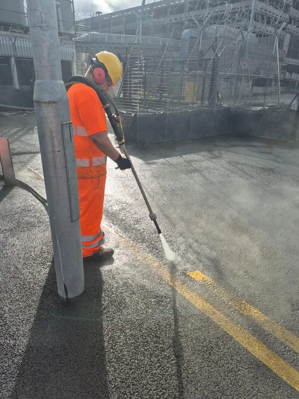 Jetting rig worker clearing a faded yellow line beside a steel post, a bright water fan cutting into the tarmac