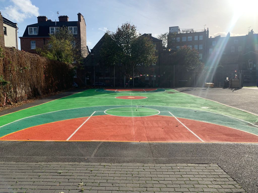 Basketball court with green, teal and orange surface coating and white line markings, overlooked by brick houses and flats