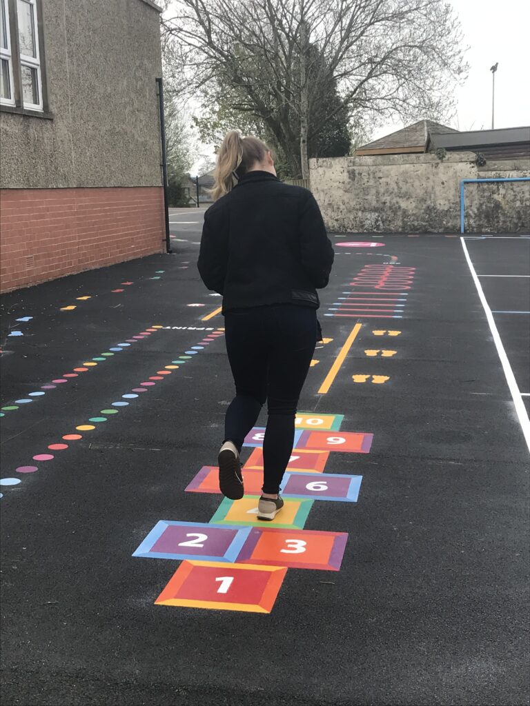 Woman in black hopping along a rainbow square hopscotch marking on new tarmac, with coloured spot trails and a sprint lane beyond