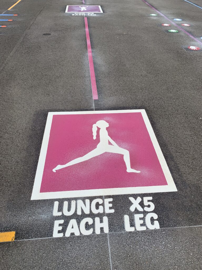 Pink fitness trail marking with a white lunge figure and the words Lunge x5 each leg, and a hop station further along