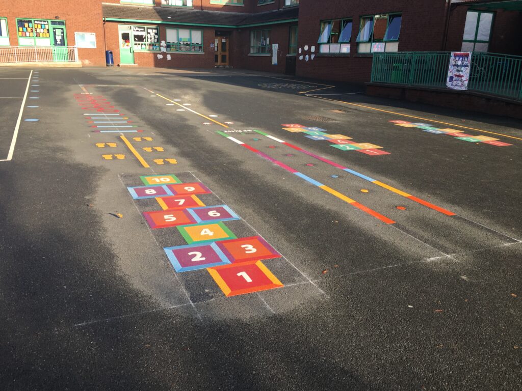 Square hopscotch, footprint trail and coloured running line markings on a playground in low sun outside a red-brick school