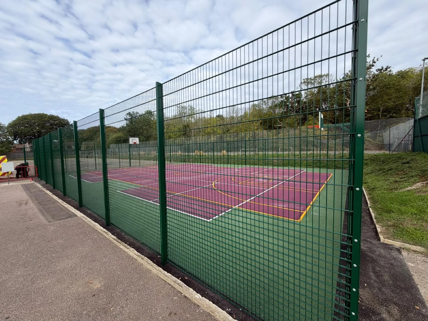 Green perimeter fencing running alongside a resurfaced maroon court with white and yellow markings, bordered by tarmac paths