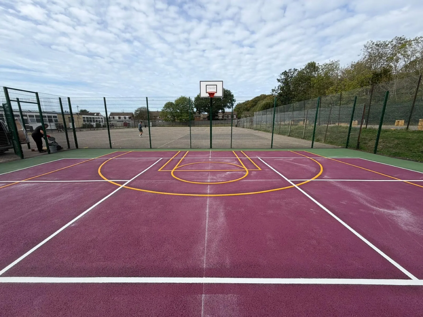 Players by a van beside a maroon multi-sport court, basketball hoop ahead and yellow and white markings underfoot